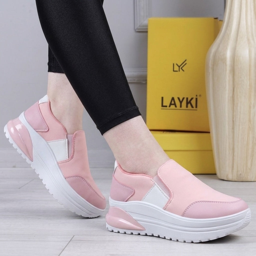 A person wearing pink slip-on platform sneakers with white soles, standing indoors next to a yellow LAYKI shoe box and a decorative vase.