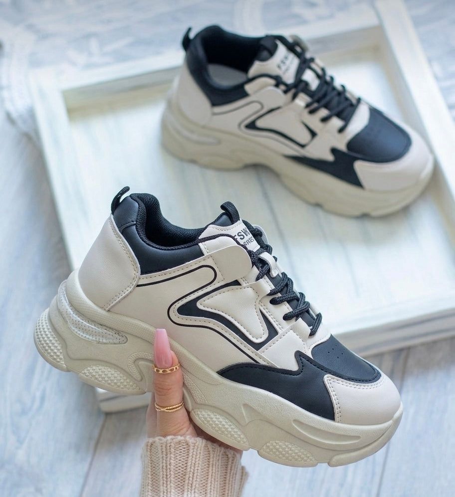 A pair of chunky platform sneakers in cream and black, with one shoe held by a hand and the other resting on a white tray in the background.