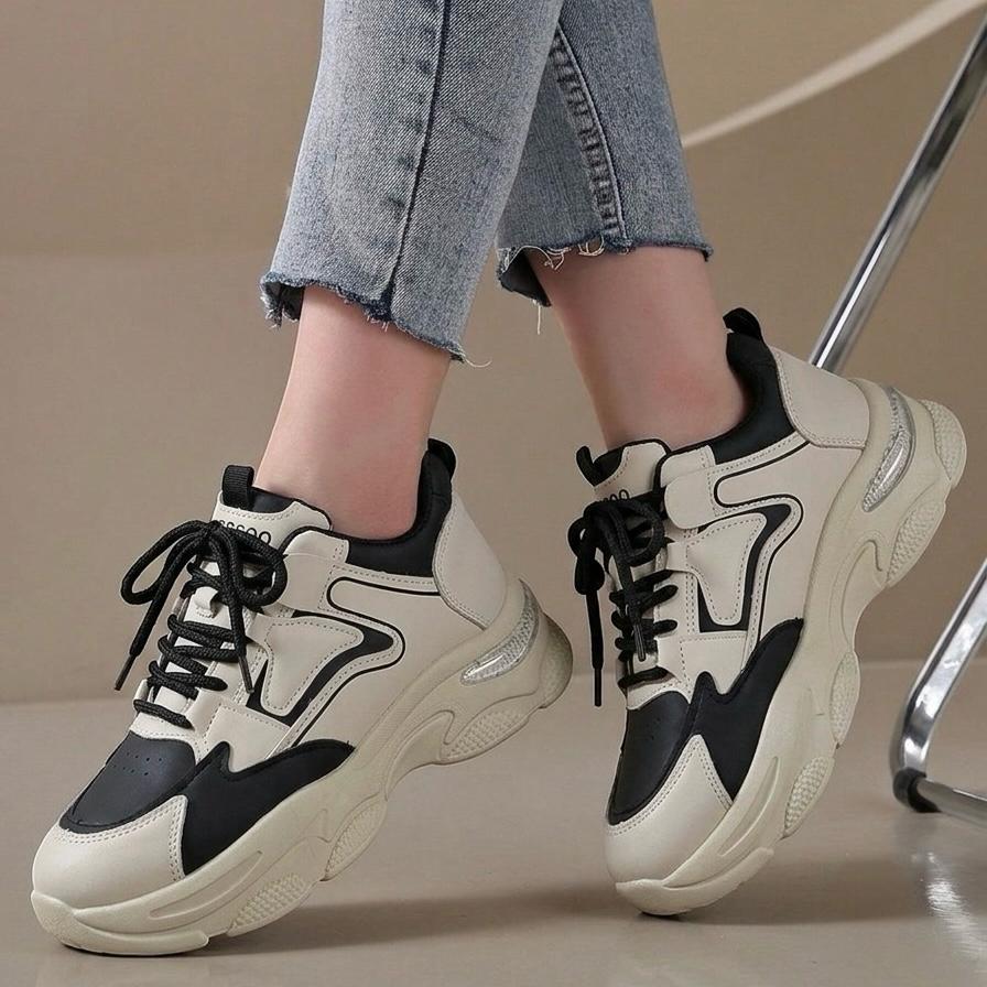 A person wearing chunky low-top sneakers in off-white and black, paired with light-wash frayed-hem jeans. The shoes feature a thick sculpted sole, black laces, and black contrast panels on a beige indoor floor.