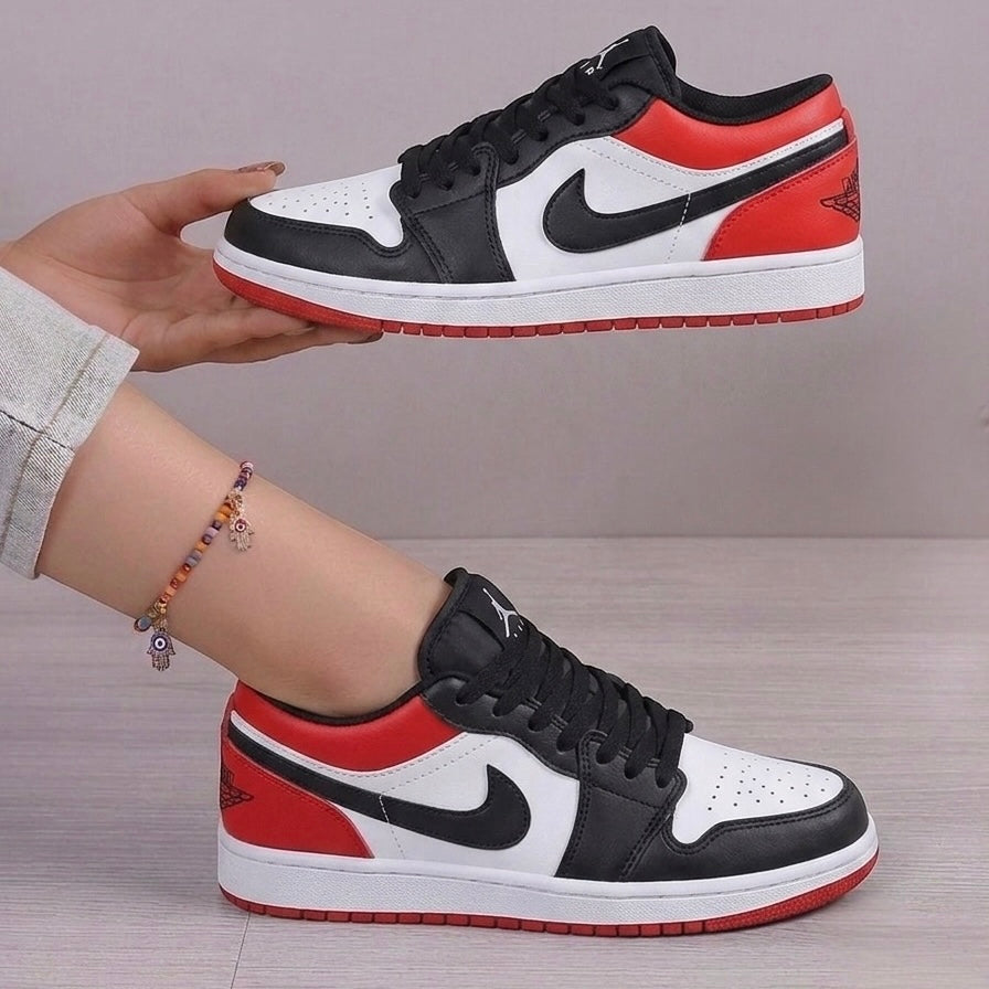 A pair of Air Jordan 1 Low sneakers in a classic black, white, and red colorway. One shoe is held up by a hand, while the other is shown worn on a foot.