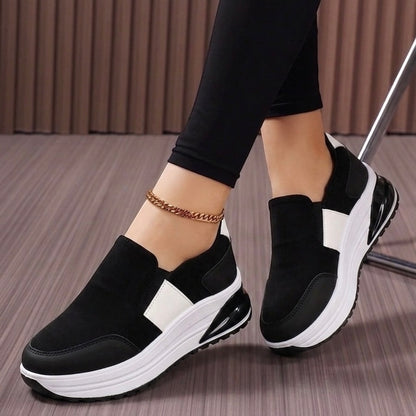 A person wearing black slip-on platform sneakers with white side panels and chunky white soles, paired with black leggings and a gold anklet, standing on a gray wood floor.