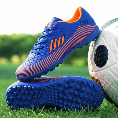 Blue and orange soccer cleats with turf studs, shown on grass next to a soccer ball. The top shoe displays the model name &