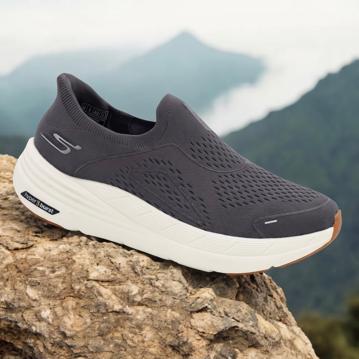 A dark gray slip-on athletic shoe with a thick white sole, resting on a large rock with a blurred mountain background.