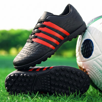 A pair of black soccer cleats with three red stripes, shown on a green grass field next to a soccer ball. One shoe is standing upright while the other lies flat to display its textured sole.