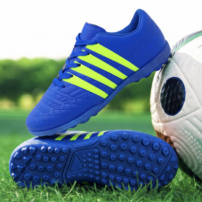 A pair of blue soccer cleats with bright neon yellow stripes, shown on a green grass field with a soccer ball partially visible on the right.