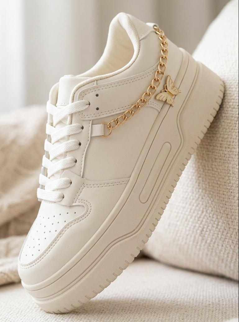 A single low-top sneaker in off-white leather with white laces and a chunky sole, styled on a soft beige textured surface with a draped fabric background. A decorative gold chain and small gold charm are attached near the collar.