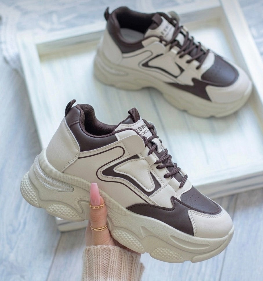 A pair of chunky platform sneakers in cream and dark brown, with one shoe being held by a hand with pink manicured nails and gold rings, and the other resting on a white wooden tray in the background.