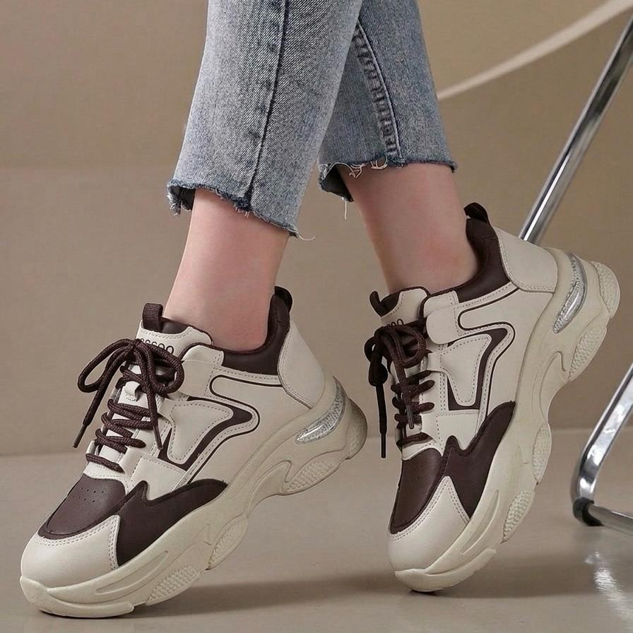 A person wearing chunky low-top sneakers in off-white and dark brown, paired with light-wash cropped jeans. The shoes feature a thick sculpted sole, dark brown laces, and dark brown trim details.
