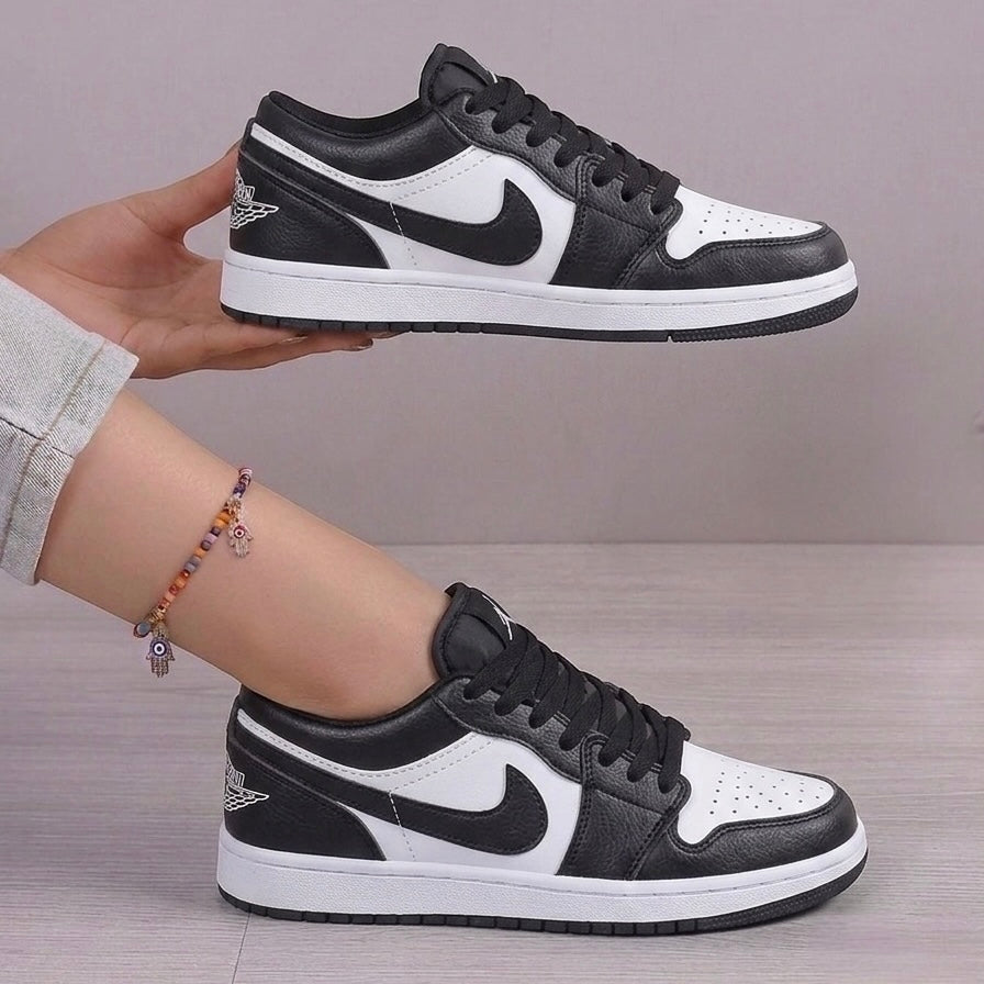 A pair of black and white Nike Air Jordan 1 Low sneakers, with one held up by a hand and the other worn on a foot, against a neutral gray background.