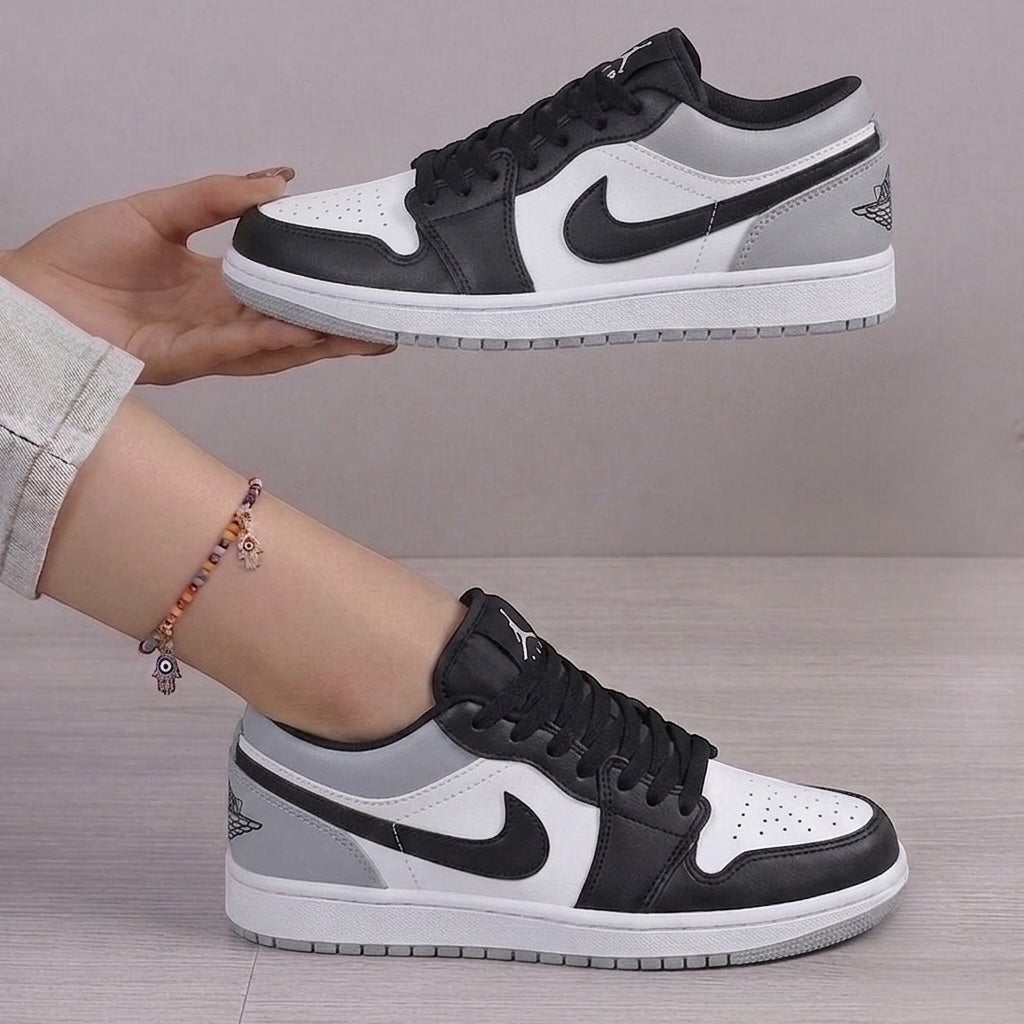 A pair of Nike Air Jordan 1 Low sneakers in white, black, and gray, with one shoe held up by a hand and the other worn on a foot, set against a neutral indoor background.