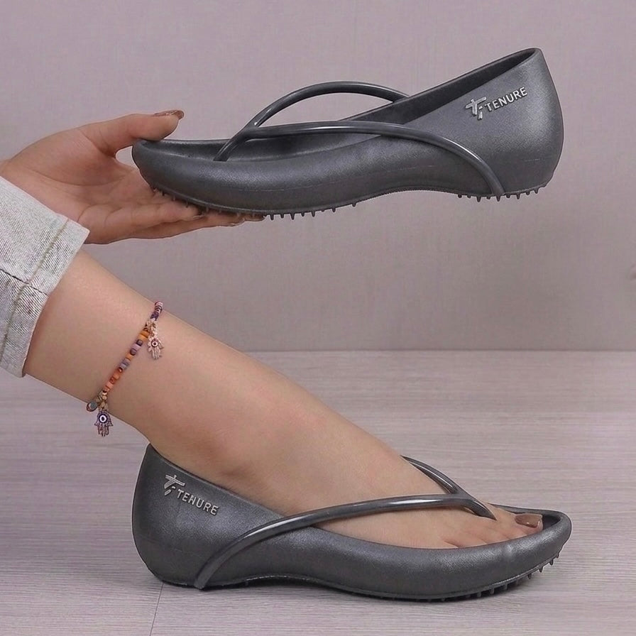 A pair of dark gray jelly slip-on shoes with metallic straps and a treaded sole, shown with one shoe on a foot and the other held by a hand against a neutral background.